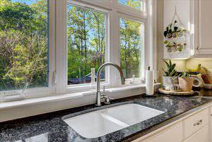 A kitchen sink with a modern faucet is set in a dark granite countertop beneath three large windows overlooking greenery. The counter has plants, a fruit basket, kitchen utensils, and paper towels.