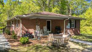 A one-story brick house with a covered porch featuring white rocking chairs and a grill. A glass patio table with four chairs is on the lawn. Trees and shrubs surround the house, and a small toy vehicle is near the porch.