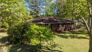 A single-story red brick house with a dark brown roof is surrounded by green trees and a grassy yard, with sunlight casting shadows across the lawn. A small pile of logs is visible on the left side.