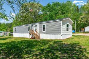A single-wide manufactured home with gray siding and white trim sits on a grassy lot. A small wooden porch with stairs leads to the front door. Trees and an American flag are visible in the background.