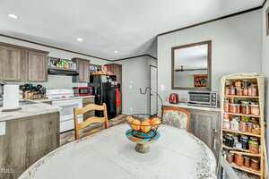 A kitchen with wood cabinets, a black refrigerator, a white oven, a red microwave, and shelves stocked with canned goods. A round dining table with a fruit bowl and wooden chairs is in the foreground.