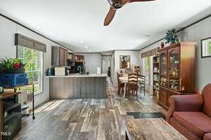 A spacious open-concept kitchen and dining area features wood-style flooring, a central island with stools, a dining table, a china cabinet with glass doors, and a red armchair in the foreground.