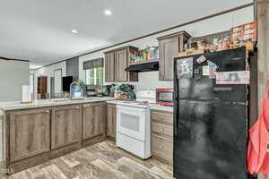 A kitchen with wood cabinets, a white stove, a black refrigerator with magnets and papers, various items on the counter, and pantry goods on top of the cabinets. The floor has a wood-look pattern.
