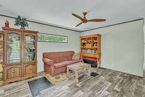 A living room with a brown sofa, a wooden coffee table, a wooden hutch, and a glass-fronted cabinet. The floor has wood-style tile, and there is a ceiling fan and a narrow window letting in daylight.
