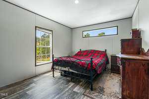 A bedroom with white walls, a large window, a single bed with a red plaid blanket, a wooden dresser, and a small wooden nightstand on a patterned rug. Sunlight enters through the windows.