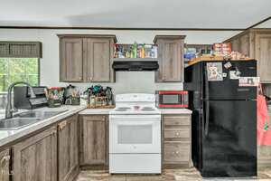A kitchen with brown cabinets, a white electric stove, a black range hood, a black refrigerator, a red microwave, and various kitchen items on the counters and above the cabinets. A window is on the left above the sink.