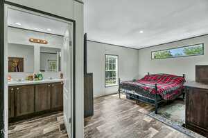 A bedroom with a metal-framed bed covered in a red plaid blanket, a wooden dresser, and a window; an adjacent bathroom with a double sink vanity and mirror is visible through an open door.
