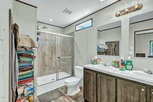 A bathroom with a glass-enclosed shower, toilet, double vanity with two mirrors, and various toiletries on the counter. A tall shelving unit on the left holds neatly folded towels and other items.