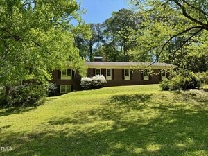 A single-story brick house with white trim sits on a grassy hill at 2118 N Woodland Road in Henderson, surrounded by green trees and bushes under a clear blue sky.