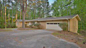 Charming single-story beige brick house at 110 Omega Road, Henderson, with a two-car garage and curved driveway, surrounded by tall pine trees and greenery. The yard features bushes and scattered pine needles.