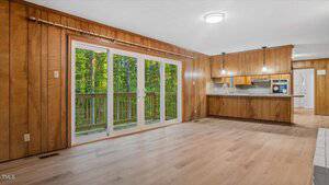 A room with wood-paneled walls, light wood flooring, and large glass sliding doors leading to a deck. The space opens into a kitchen with wood cabinets and pendant lights above the counter.