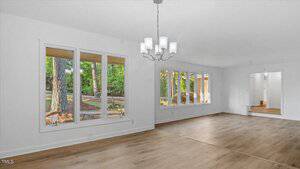 A spacious, empty living room with light wood flooring, white walls, large windows showing a wooded yard, and a modern white chandelier hanging from the ceiling.