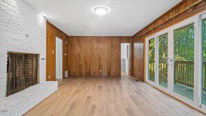A room with wood-paneled walls, light wood flooring, a white brick fireplace on the left, sliding glass doors on the right, and a view of a deck and trees outside. A small door leads to another room.