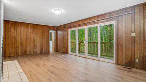 A room with wood-paneled walls, light wood flooring, and a set of glass double doors leading to a deck with a view of trees outside. A doorway on the left leads to another room. Ceiling lights are on.
