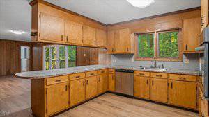 A kitchen with wooden cabinets, a double sink under two windows, a dishwasher, and marble-pattern countertops. The space opens to a room with wood-paneled walls and a large glass door leading outside.
