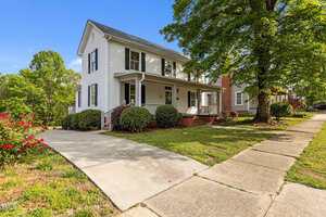 A two-story white house at 116 W Front Street in Oxford, featuring a covered front porch, black roof, and driveway, surrounded by green lawn, bushes, a tree, and a sidewalk under a clear blue sky.