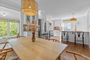 A modern kitchen and dining area with light wood furniture, stainless steel appliances, shiplap walls, and pendant lighting. Large windows bring in natural light, and a vase with dried plants sits on the dining table.