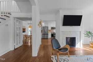 A modern living space with hardwood floors, a black chair on a rug, a white brick fireplace with a mounted TV, and a view into a dining area and kitchen with stainless steel appliances. A staircase is visible on the left.