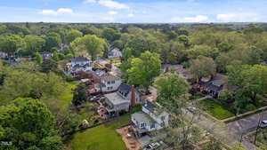 Aerial view of a suburban neighborhood with tree-lined streets, detached houses, green lawns, and cars parked along the roads on a clear day. Dense trees surround and fill the area.
