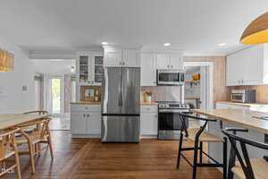 A modern kitchen with white cabinets, stainless steel appliances, wood countertops, and a dining area with wooden chairs. The space is bright with ample natural light and a mix of recessed and pendant lighting.
