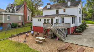 A two-story white house with a red foundation features a covered back porch, wooden stairs, and a gravel patio area with seating and a fire pit, alongside a large driveway and neighboring homes.