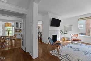 A bright living room with wood floors, a patterned rug, a modern blue chair, and a fireplace with a TV above it. Large windows let in natural light. A dining area with a wooden table is visible through an open doorway.