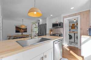 A modern kitchen with white cabinets, light wood countertops, stainless steel appliances, and a gooseneck faucet. An open dining area and a laundry room with washer and dryer are visible in the background.