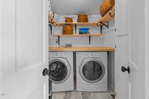 A laundry room with a front-loading washer and dryer beneath wooden shelves holding baskets and decorative items. The floor is tiled, and the space is enclosed by white doors.