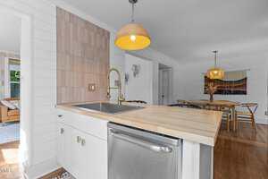A modern kitchen with a light wood countertop, stainless steel sink, gooseneck faucet, and dishwasher. The backsplash features neutral tiles, and a round pendant light hangs above. A dining area is visible in the background.