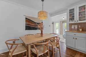 A dining area with a light wooden table, six matching chairs, a woven pendant light, wall art, and built-in cabinets. Dried flowers in a vase are on the table, and double doors lead to a balcony outside.