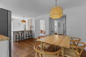 A dining area with a wooden table and six chairs sits under a woven pendant light; in the background, a kitchen island with two stools, wood floors, and white shiplap walls are visible.