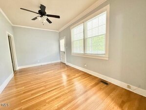 A bright, empty room with light gray walls, hardwood floors, white trim, a ceiling fan with lights, large windows with blinds, and a door leading outside.