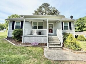 A small, single-story house at 309 19th Street in Butner, featuring light-colored siding, black shutters, a covered front porch with white railings, and concrete steps leading up to the door. Shrubs and grass surround the home.