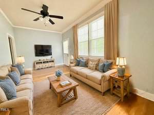 A living room with light blue walls, two beige sofas with blue cushions, a wooden coffee table, a TV mounted on the wall, a ceiling fan, and large windows with beige curtains letting in natural light.