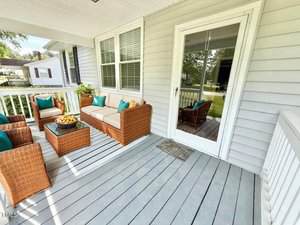A covered porch with light gray wooden flooring and white railings, featuring wicker patio furniture with teal cushions, a glass-top coffee table, and a doormat in front of a glass door leading inside.