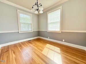 Empty room with light gray walls, white trim, wood floor, two windows with blinds, and a ceiling light fixture with four frosted glass shades. Sunlight streams through one window, casting shadows on the floor.