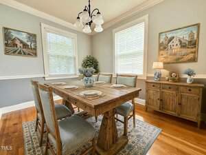 A dining room with a wooden table set for six, matching chairs with blue cushions, a sideboard, two landscape paintings, a floral rug, and light blue-gray walls. The room has hardwood floors and large windows with blinds.