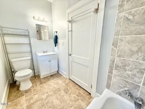 A bathroom with beige tile flooring, a white toilet, a white vanity with sink and mirror above, a metal shelving unit, a door, and a bathtub with gray tile surround. A blue towel hangs on a hook.