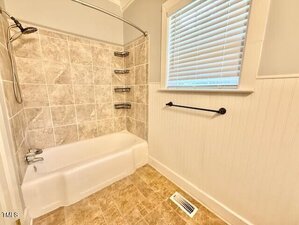 A bathroom with a white bathtub and shower, beige tile walls, three corner shelves, a window with white blinds, a black towel bar, beige tile floor, and a floor vent.