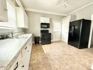 A spacious kitchen with light-colored countertops, white cabinets, a black oven and microwave, a black refrigerator, and a double sink under large windows. The floor has beige tiles and the walls are light gray.