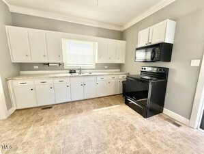 A kitchen with light gray walls, white cabinets, a black electric stove with a matching microwave above it, a double sink under a window, and beige tile flooring. The kitchen is empty with no visible furnishings.