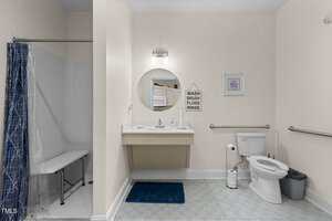 Accessible bathroom with a walk-in shower and foldable bench, a floating sink with a mirror, a framed print, a toilet with grab bars, waste bin, and a blue bath mat on patterned flooring.
