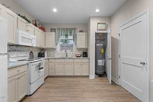 A kitchen with light-colored cabinets, white appliances, a tile backsplash, and wood flooring. A window with a green curtain is above the sink, and a water heater is visible in a corner closet with the door open.
