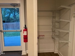 A pantry with white wire shelving is shown next to a glass exit door. A red fire extinguisher is mounted on the wall near the door. The area outside the door includes a lawn, a fence, and trees.