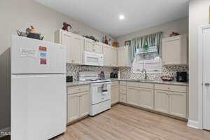 A kitchen with cream-colored cabinets, white appliances including a refrigerator, stove, and microwave, light tile backsplash, wood-look flooring, and a window with green curtains above the sink.