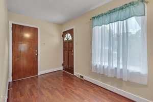 A small entryway with light yellow walls, two wooden doors, wood flooring, and a window with a blue and white curtain on the right side.