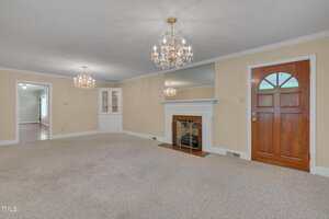 A spacious living room with beige carpet, a wooden door, a white fireplace with a mirror above it, two crystal chandeliers, built-in corner shelves, and cream-colored walls.