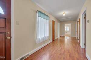 A hallway with light yellow walls, wooden flooring, a window with a sheer white curtain and blue valance, multiple doors on the right, and a ceiling light fixture. The entry door is partially visible on the left.