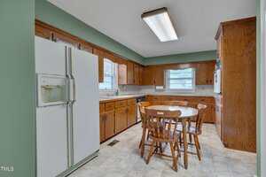 A kitchen with wood cabinets, a white refrigerator, a round wooden table with four chairs, laminate countertops, a fluorescent ceiling light, and two windows letting in natural light.