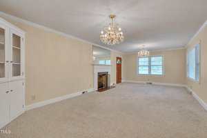 A spacious living room with beige walls and carpet, a white fireplace with a mirror above it, two chandeliers, several windows, and a built-in corner cabinet. The front door is visible in the background.